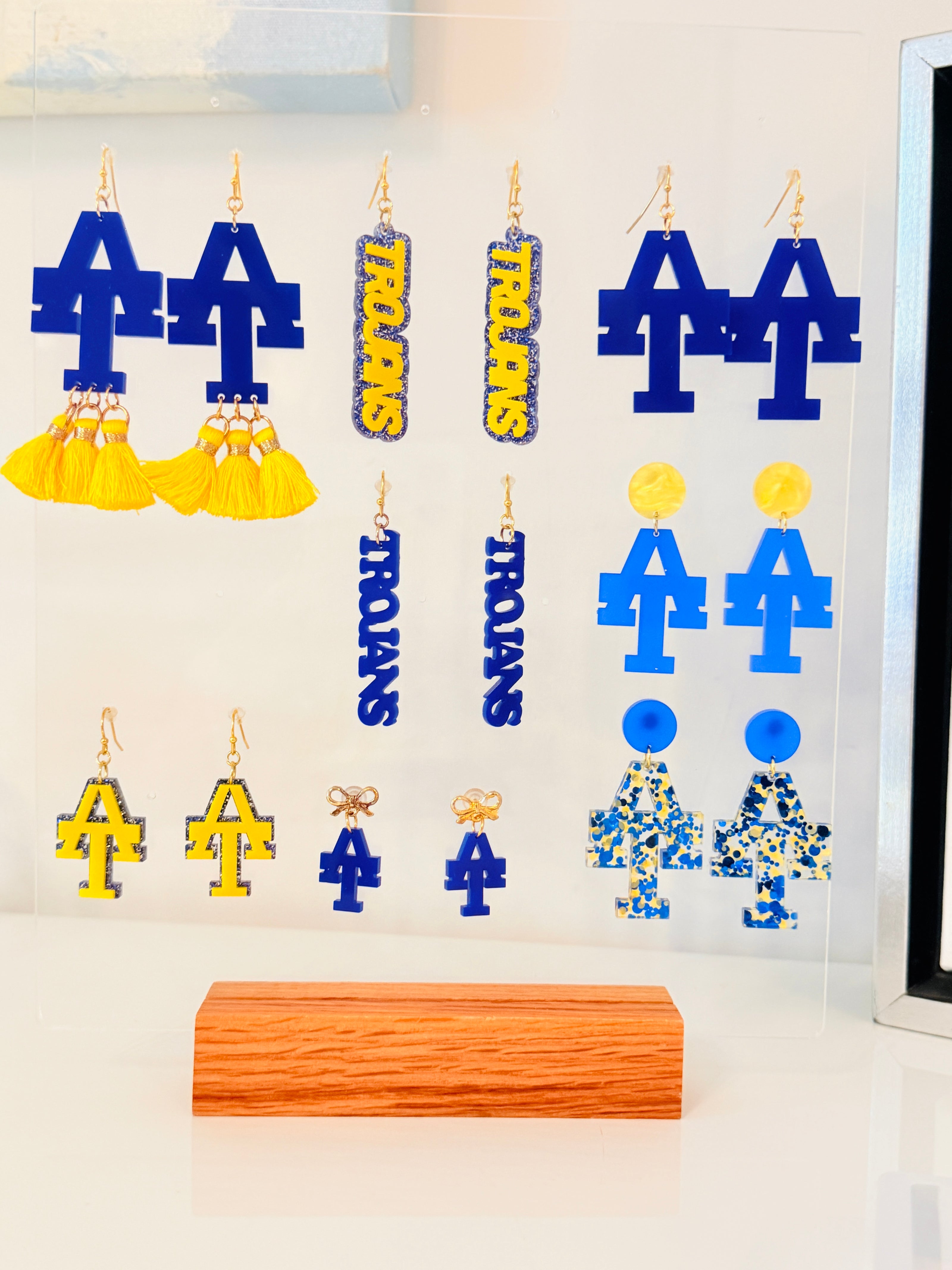 A set of blue and yellow Louisiana Tech Troyan themed earrings displayed on a clear acrylic stand, with a wooden block base. The earrings include large 'T' and 'A' shapes, 'TROYAN' nameplates, and patterned figures.