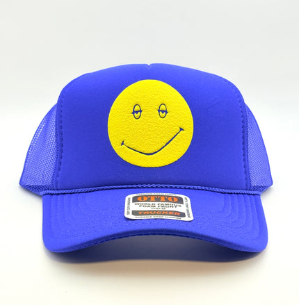 A royal blue trucker hat with a yellow smiley face patch on the front panel. The hat features a structured blue front panel and curved brim, with blue mesh side and back panels.