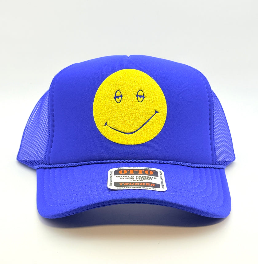 A royal blue trucker hat with a yellow smiley face patch on the front panel. The hat features a structured blue front panel and curved brim, with blue mesh side and back panels.