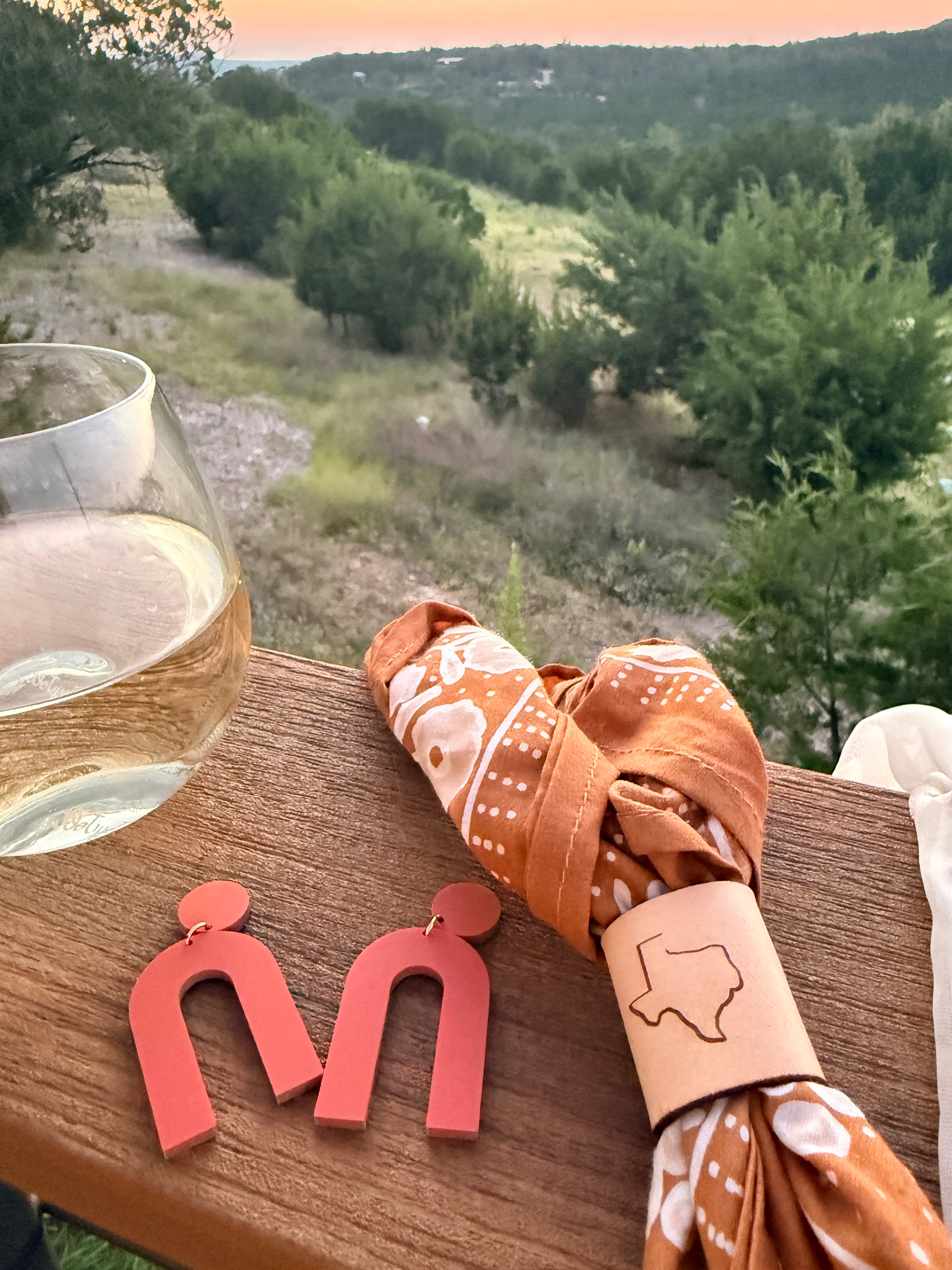 A close-up lifestyle shot of a rustic wooden table set outdoors with a scenic hillside landscape in the background. The table features a rolled terracotta orange and white patterned bandana napkin with a tan leather holder engraved with the outline of Texas, alongside a glass of white wine and a pair of coral-red wooden letter 'M' earrings.