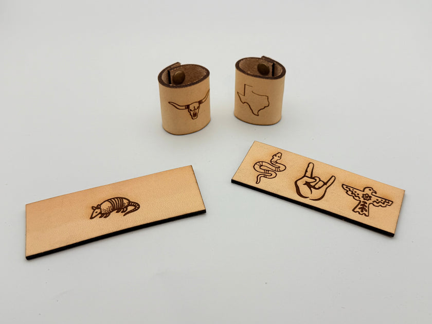 Four natural leather engraved accessories on a white background: two cylindrical rings with bull and Texas state designs, and two rectangular bars with armadillo, snake, hand, and bird designs.