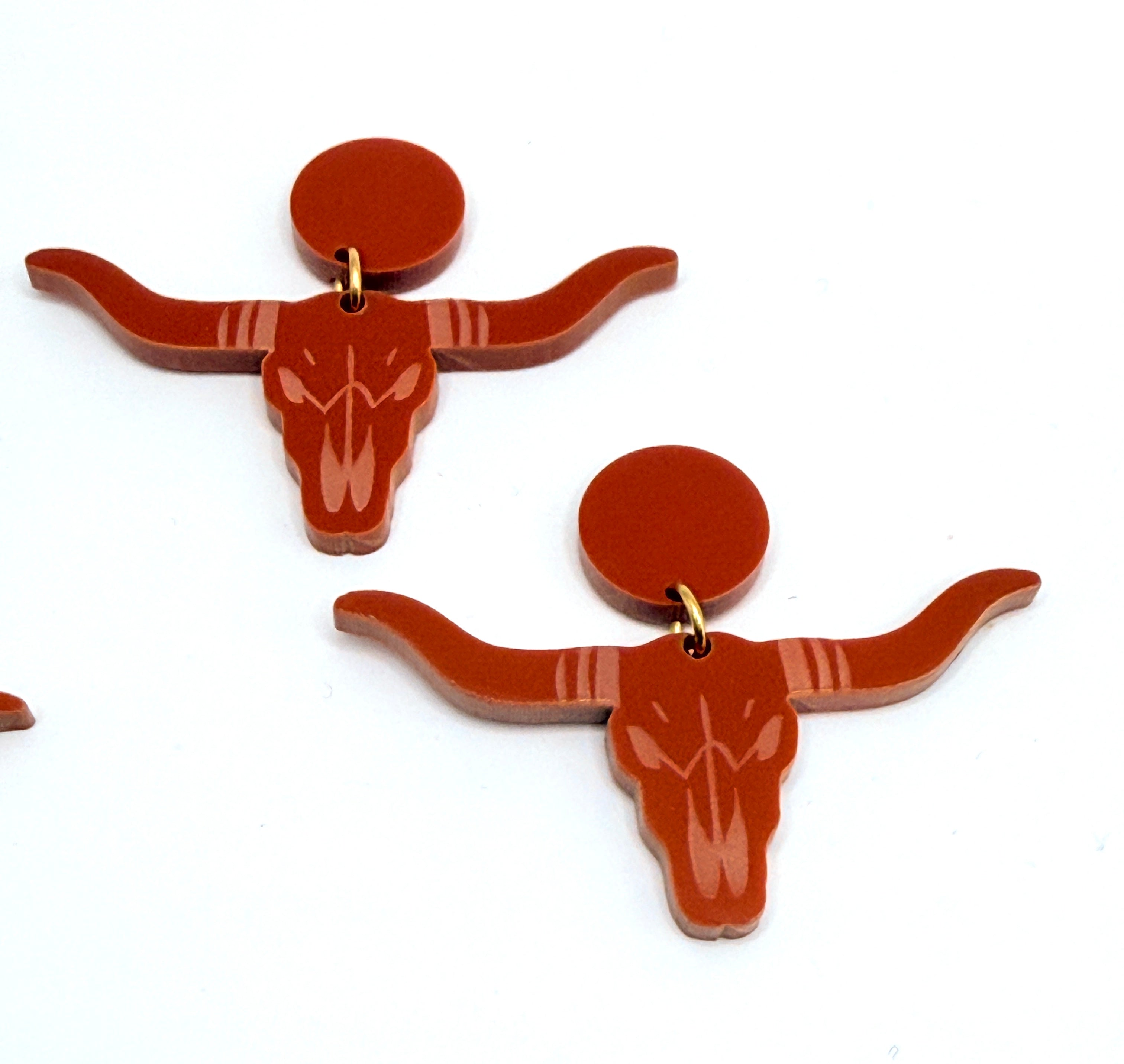 A pair of burnt-orange acrylic longhorn skull earrings with circular backings, displayed on a plain white background.