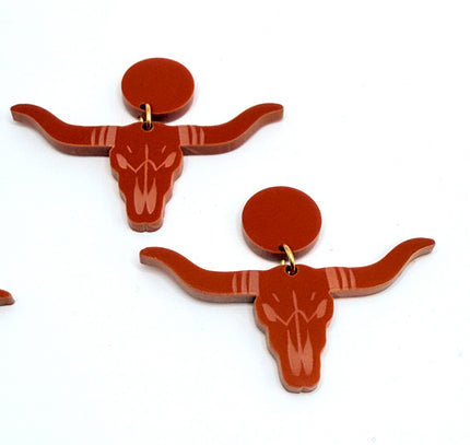 A pair of burnt-orange acrylic longhorn skull earrings with circular backings, displayed on a plain white background.