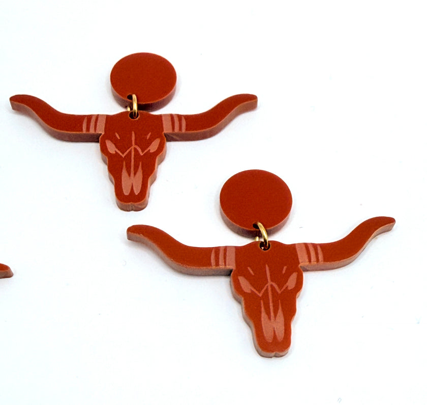 A pair of burnt-orange acrylic longhorn skull earrings with circular backings, displayed on a plain white background.