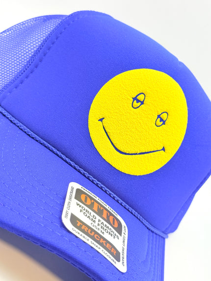 A close-up of a bright blue trucker hat featuring a large yellow smiley face patch on the front panel and a visible OTTO World Famous Foam Mounts Trucker label on the side.