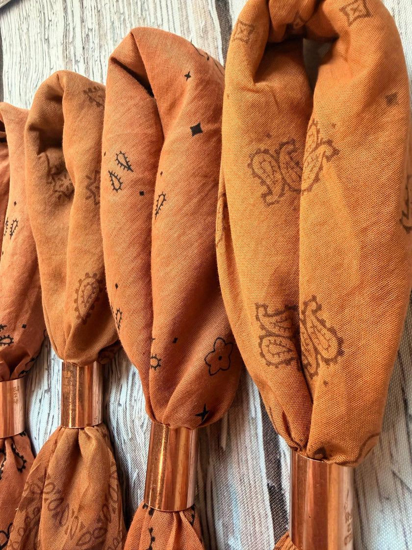 A close-up of several rust-orange fabric napkins or bandanas tied around copper-colored handles, arranged vertically against a whitewashed wood background.