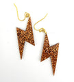 A pair of gold-tone fishhook earrings with lightning bolt-shaped drops covered in sparkling copper glitter, shown on a plain white background.