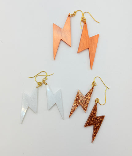 Four lightning bolt dangle earrings arranged on a plain white background. The set includes two copper-toned matte dangles with gold hooks, two silver-toned matte dangles with gold hooks, and two copper-toned glitter dangles with gold hooks.