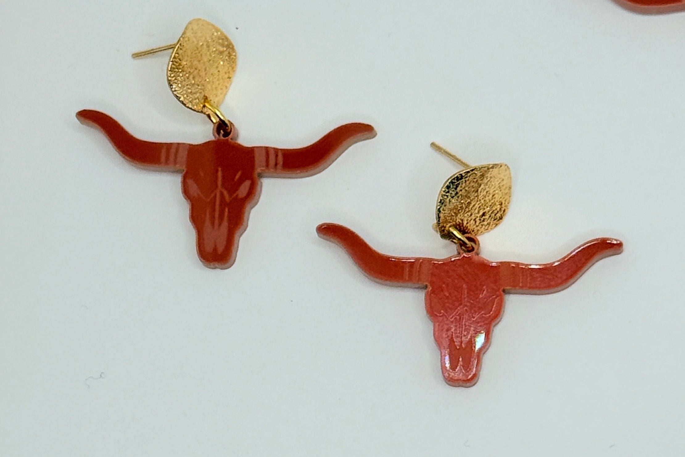 A pair of bohemian-style earrings featuring reddish-brown longhorn motifs with gold studs, alongside partial matching earrings with iridescent white circular discs on a white background.