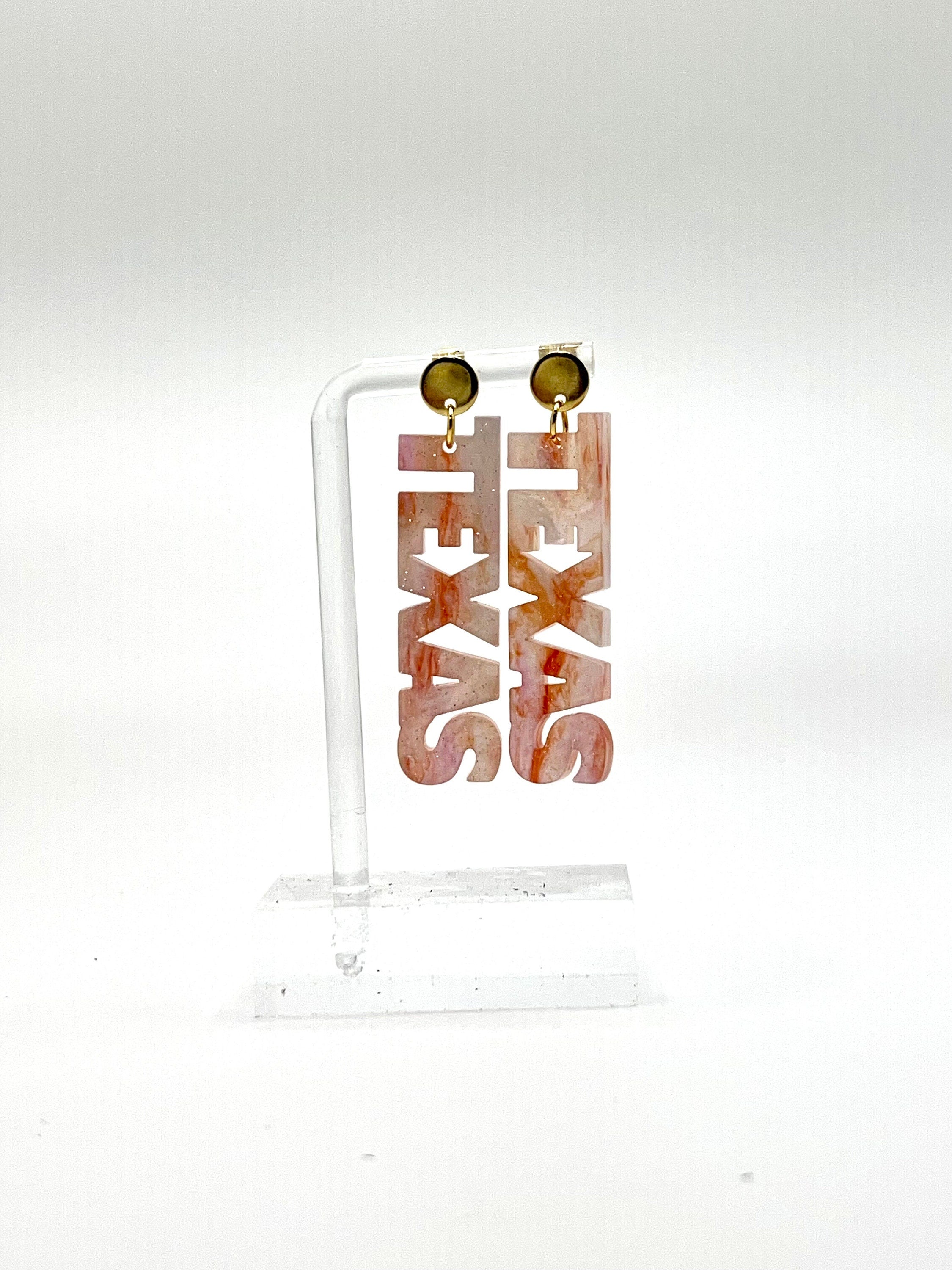 A pair of acrylic earrings in a marble pattern spelling out 'TEXAS' in a vertical orientation, displayed on a clear stand against a white background.