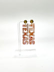 A pair of acrylic earrings in a marble pattern spelling out 'TEXAS' in a vertical orientation, displayed on a clear stand against a white background.