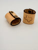 Two tan leather ring stands on a plain white background, one standing upright with a dark brown edge and a Texas state outline engraving, and the other lying on its side with two metal snap buttons.