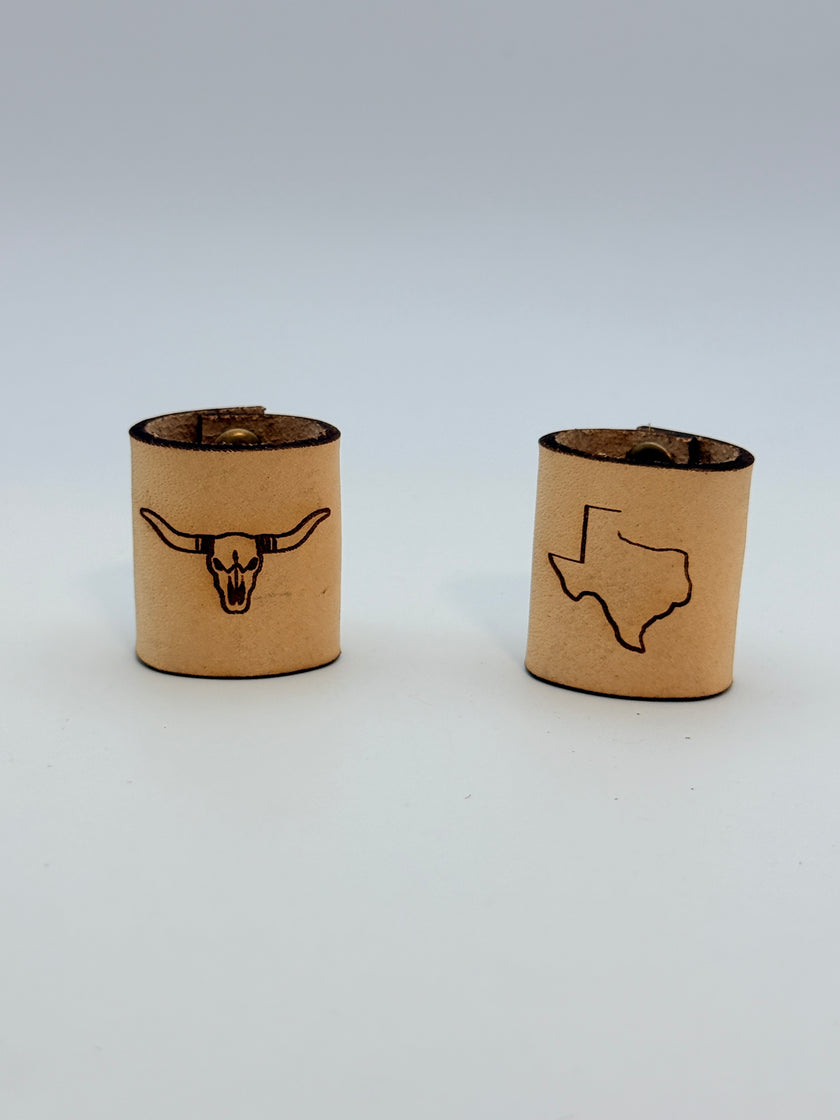 Two tan cylindrical bandana slides with engraved black designs, one featuring a longhorn skull and the other the outline of Texas, on a light gray background.