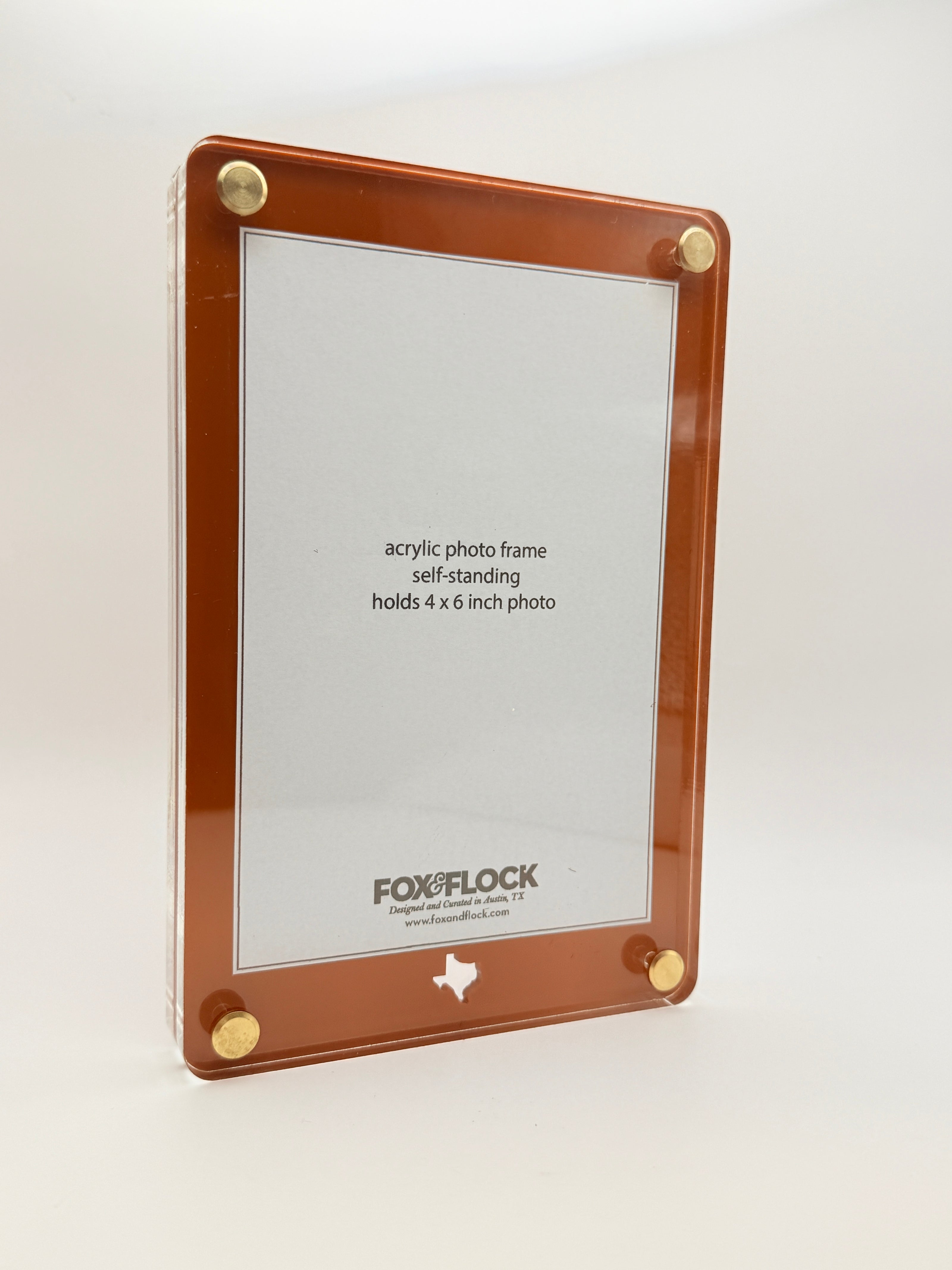 A Fox & Flock self-standing acrylic photo frame with a copper-brown border and gold metal corner hardware, shown on a plain white background.