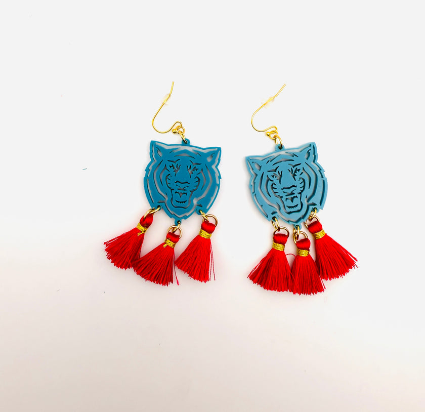 A pair of bright pink acrylic earrings in the shape of a tiger's head, attached to gold-colored hooks, displayed against a light background with a red tassel in the corner.