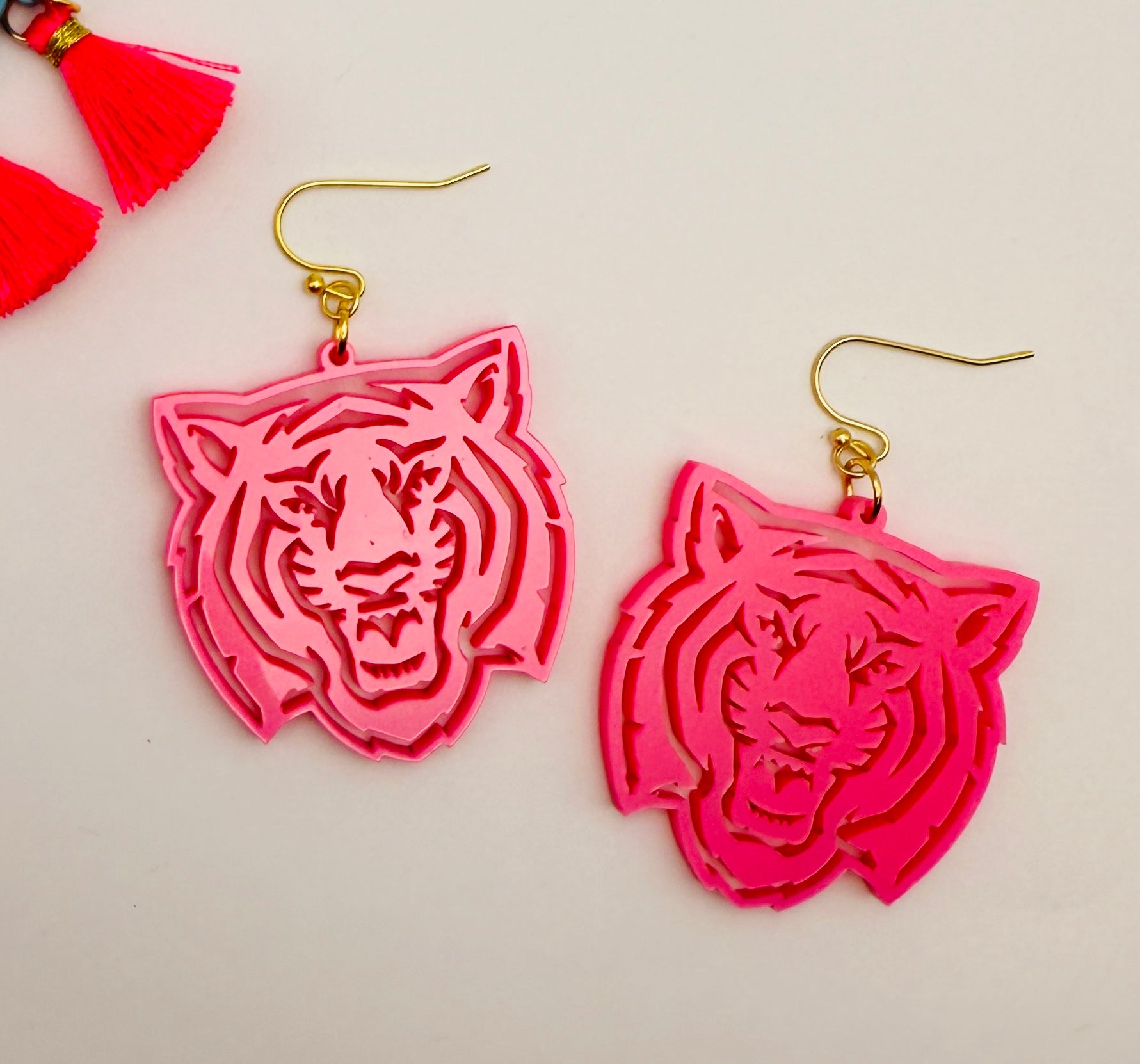 A pair of bright pink acrylic earrings in the shape of a tiger's head, attached to gold-colored hooks, displayed against a light background with a red tassel in the corner.