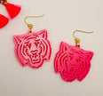 A pair of bright pink acrylic earrings in the shape of a tiger's head, attached to gold-colored hooks, displayed against a light background with a red tassel in the corner.