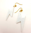 A pair of white lightning bolt dangle earrings with gold fishhook wires, displayed on a clean white background.
