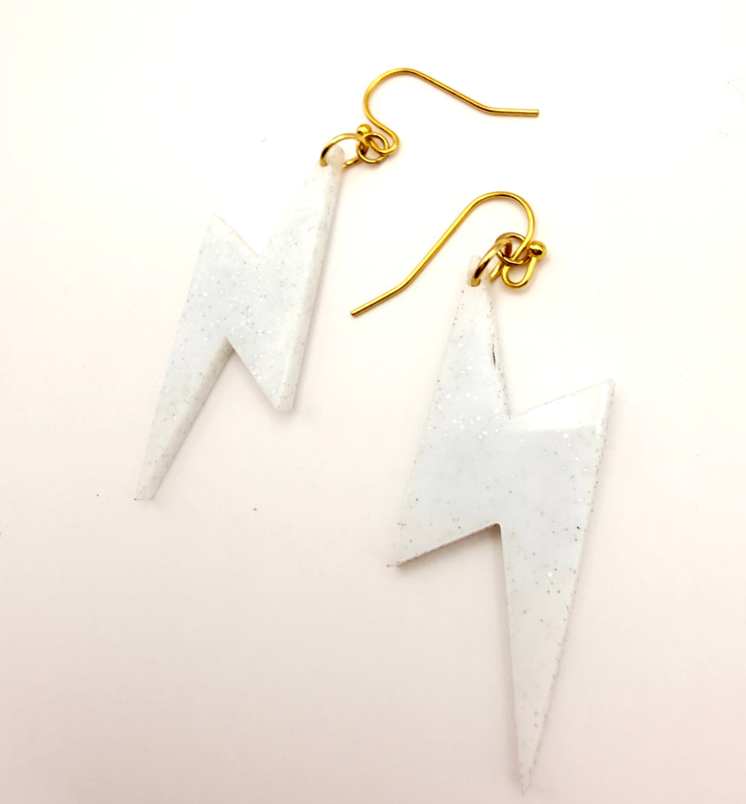 A pair of white lightning bolt dangle earrings with gold fishhook wires, displayed on a clean white background.