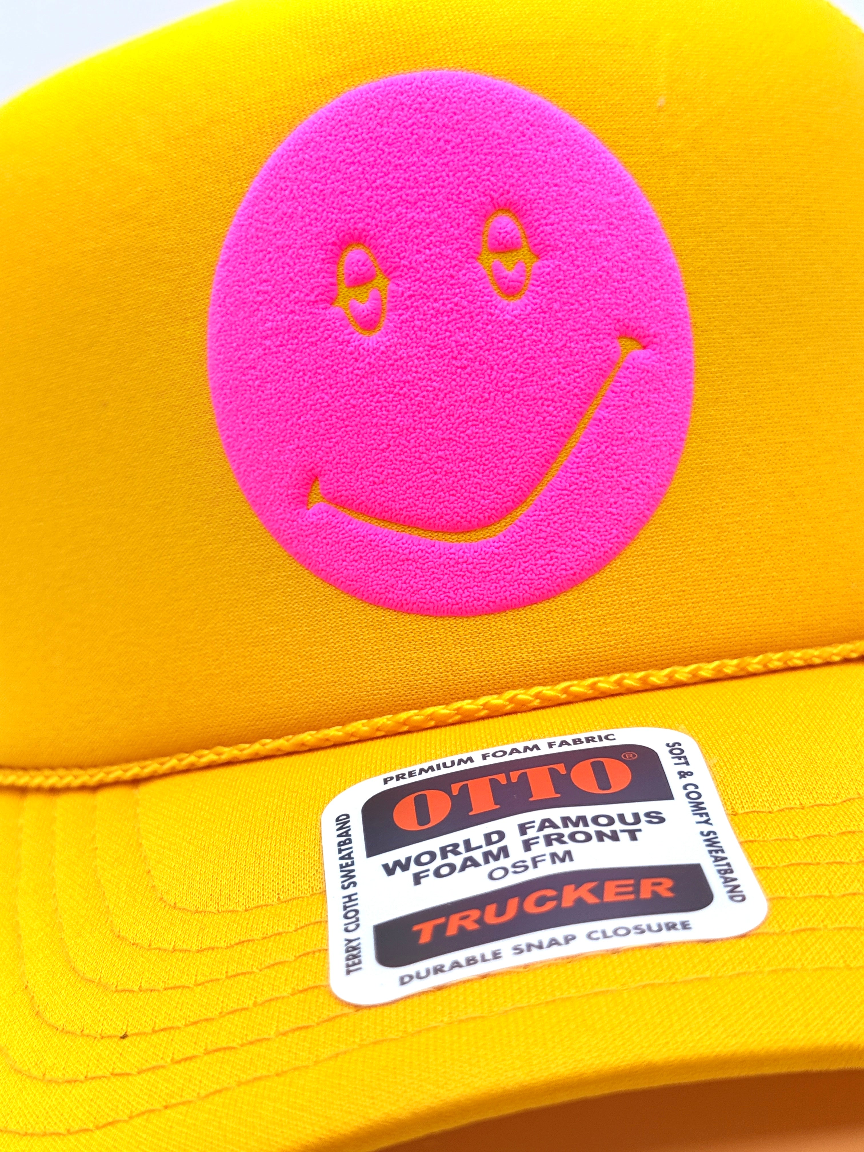Close-up of a bright yellow trucker cap with a large pink foam smiley face patch on the front and a visible OTTO TRUCKER sticker on the brim.