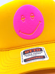 Close-up of a bright yellow trucker cap with a large pink foam smiley face patch on the front and a visible OTTO TRUCKER sticker on the brim.