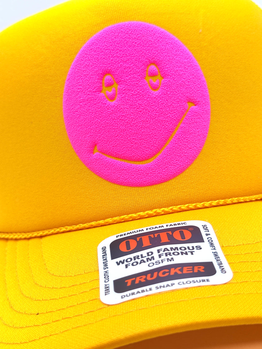 Close-up of a bright yellow trucker cap with a large pink foam smiley face patch on the front and a visible OTTO TRUCKER sticker on the brim.