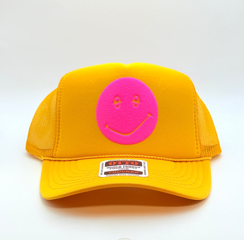 A yellow trucker hat with a bright pink smiley face patch on the front, photographed against a plain white background.