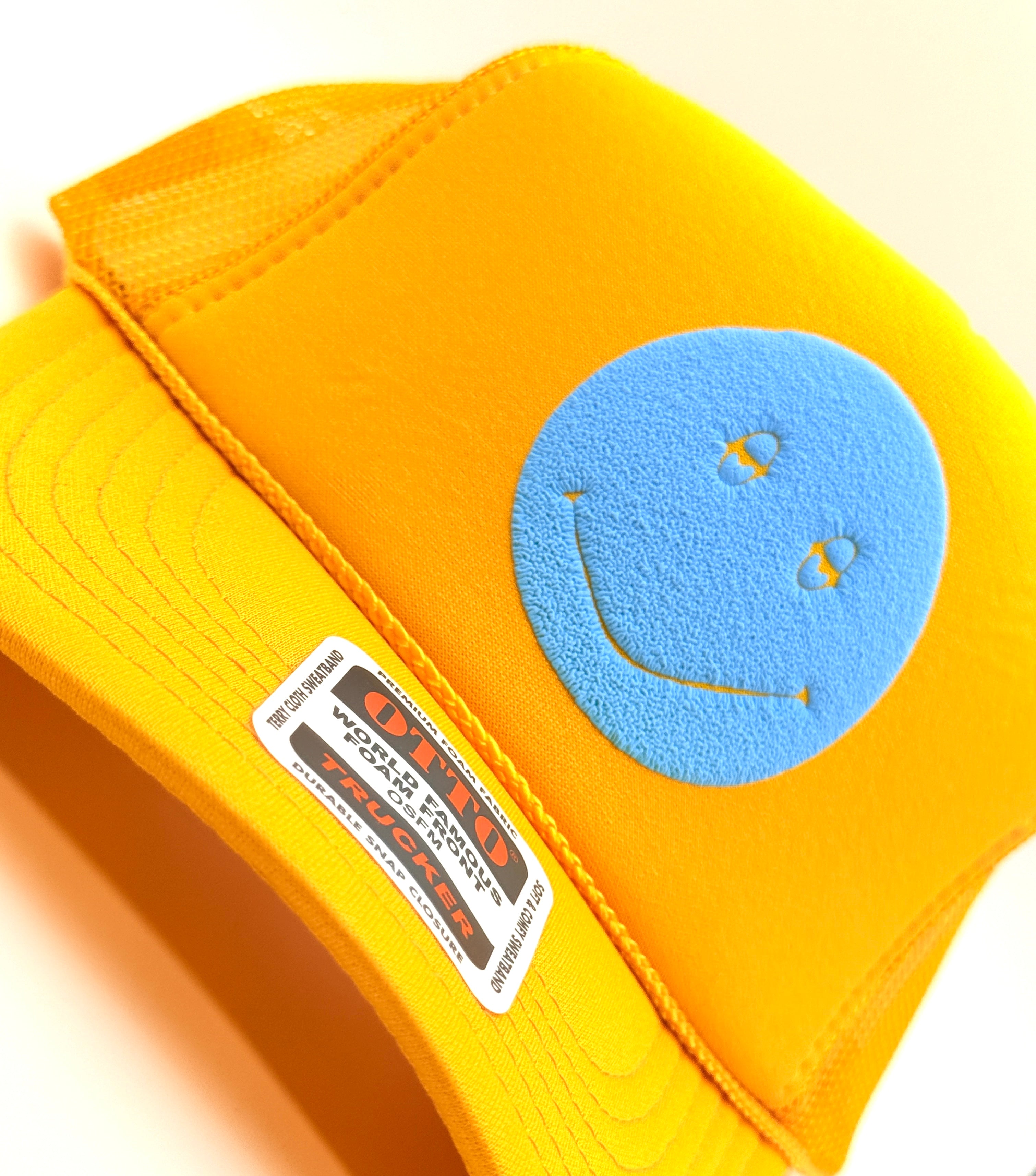 A close-up of a bright yellow trucker hat featuring a blue smiley face patch on the front and a visible 'Gildan' brand label on the brim.