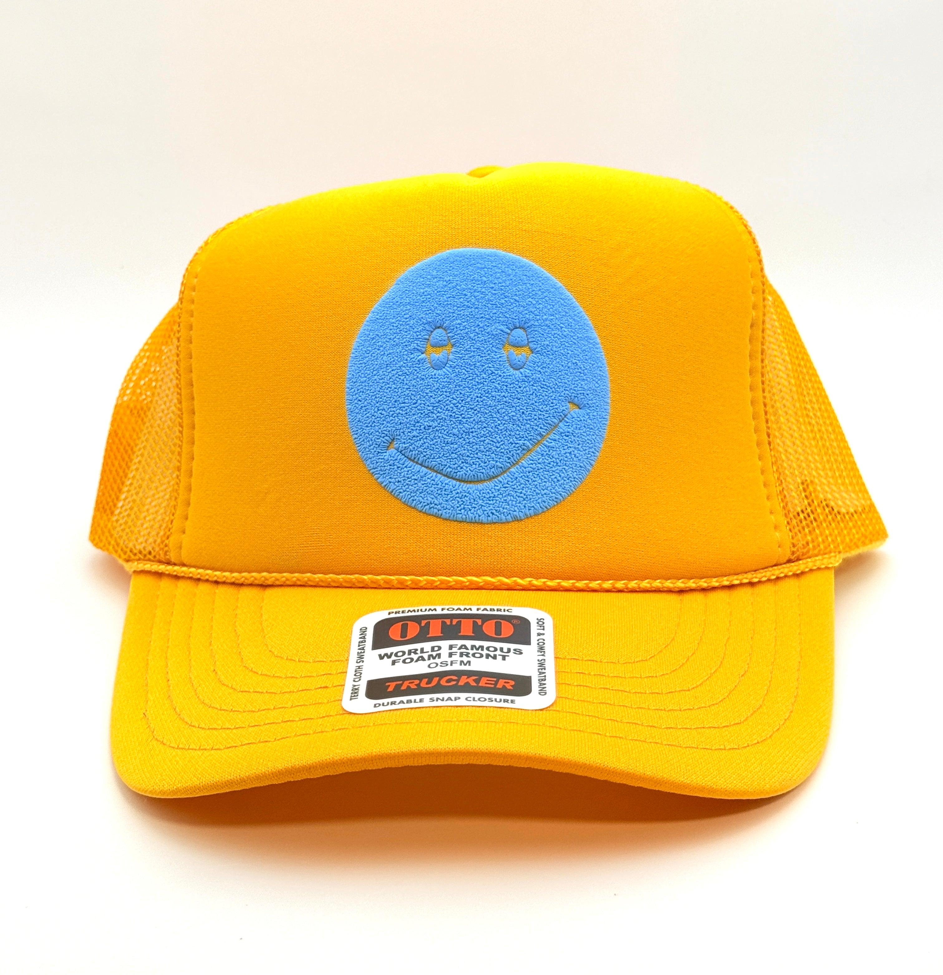 A yellow trucker hat with a blue smiley face patch on the front and a visible OTTO World Family Trading Company trucker label on the brim, photographed on a clean white background.
