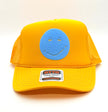 A yellow trucker hat with a blue smiley face patch on the front and a visible OTTO World Family Trading Company trucker label on the brim, photographed on a clean white background.