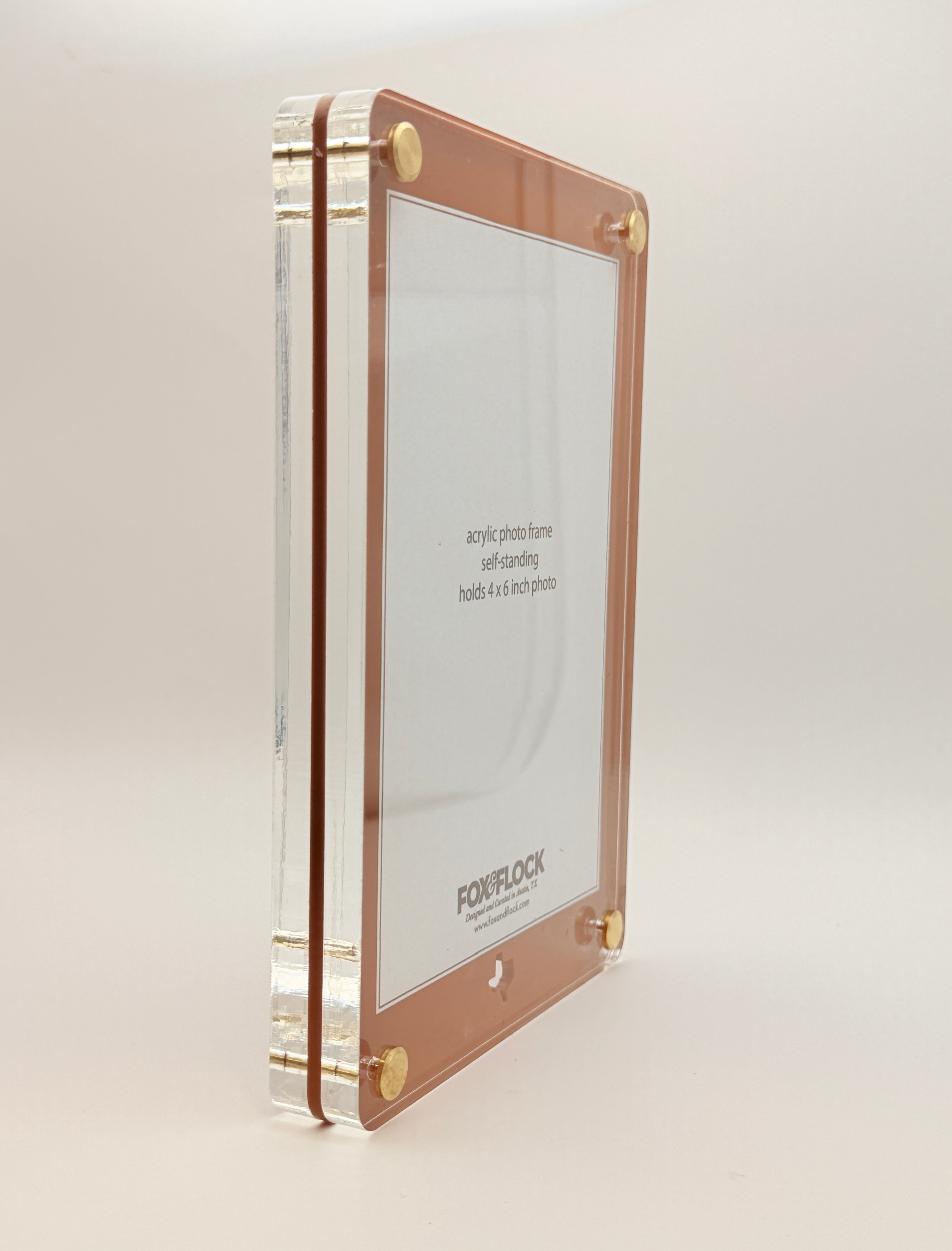 A vertical acrylic picture frame with a rose gold border and gold corner hardware, shown standing upright on a light beige background. The clear acrylic side panels are visible, and the inside insert displays the brand name 'FOX and FLOCK'.