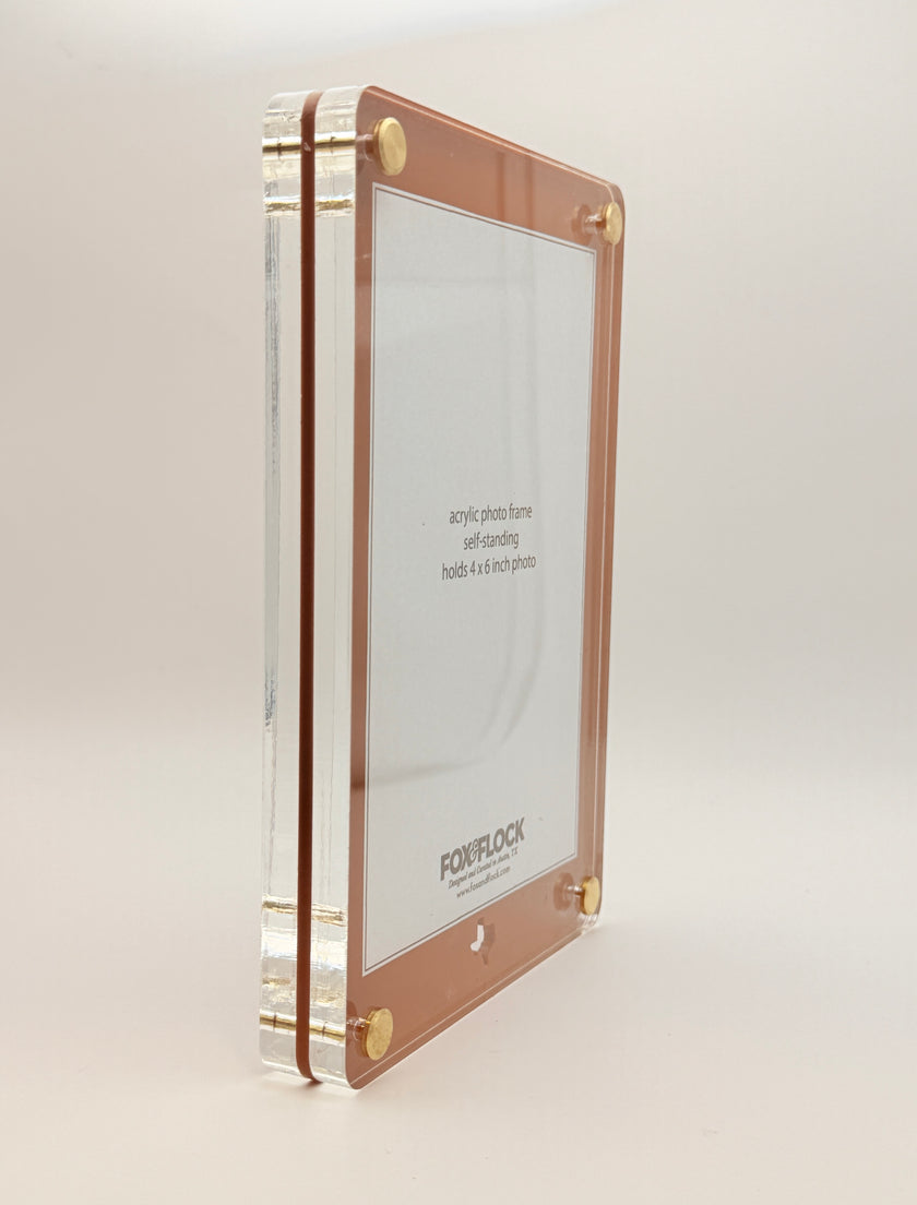 A vertical acrylic picture frame with a rose gold border and gold corner hardware, shown standing upright on a light beige background. The clear acrylic side panels are visible, and the inside insert displays the brand name 'FOX and FLOCK'.