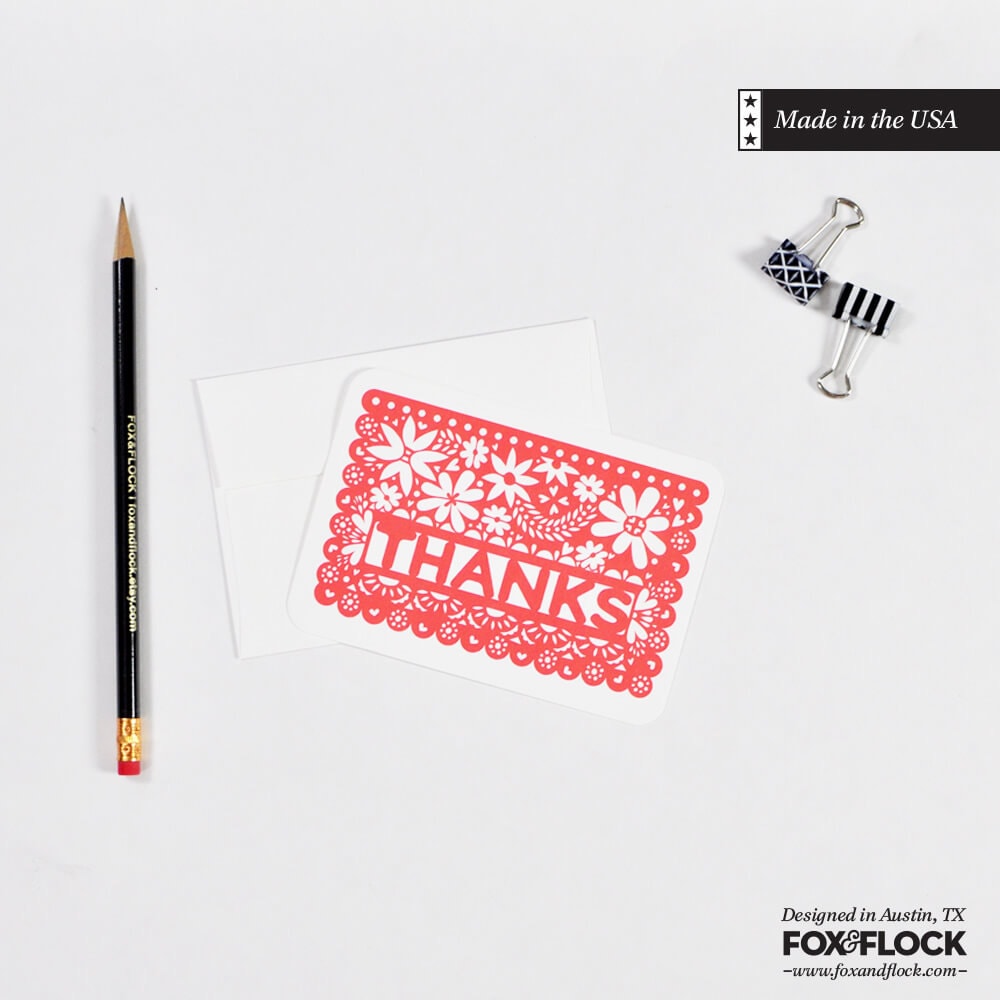A flat notecard with a red hand-drawn floral design on it, labeled 'THANK', accompanied by an envelope and a decorative clip on a white background. The notecard is presented on a made-in-USA label.