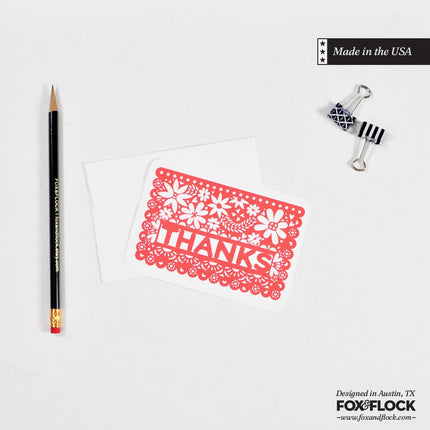 A flat notecard with a red hand-drawn floral design on it, labeled 'THANK', accompanied by an envelope and a decorative clip on a white background. The notecard is presented on a made-in-USA label.